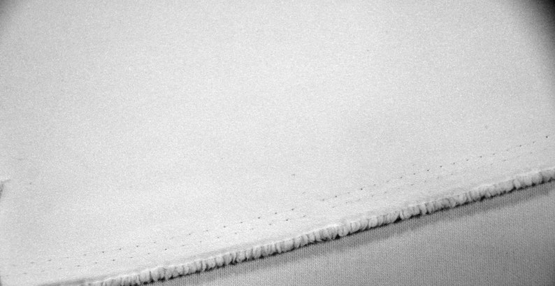 以下が含まれることがあります： テクスチャ加工された表面を持つ白い生地のクローズアップ。端はほつれており、端の近くにステッチの線が見えます。生地は、おそらく綿またはリネンである軽い素材のようです。