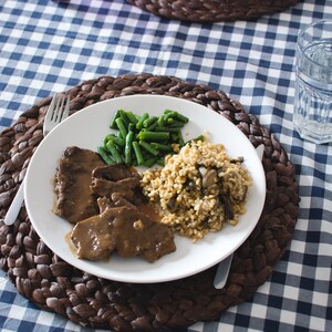 May include: A white plate with a brown sauce, sliced meat, green beans, and a side of bulgur wheat with mushrooms. The plate is on a brown wicker placemat on a blue and white checkered tablecloth.