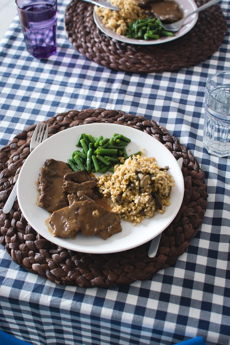 May include: A white plate with a brown sauce, sliced meat, green beans, and a side of bulgur wheat with mushrooms. The plate is on a brown wicker placemat on a blue and white checkered tablecloth.