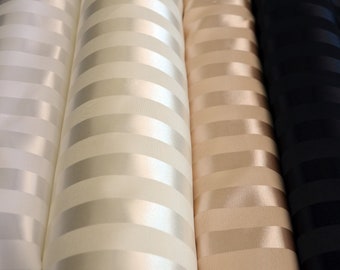 Puede incluir: Tres rollos de tejido de satén a rayas en blanco, beige y negro. El tejido tiene un brillo sutil y una textura suave.