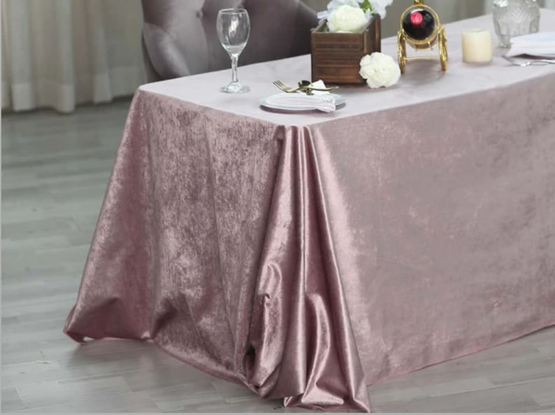 May include: A dusty rose velvet tablecloth draped over a table with a white plate, cutlery, and a glass of water. The tablecloth has a soft, luxurious feel and is perfect for adding a touch of elegance to any event.
