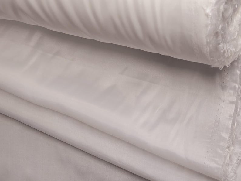 Puede incluir: Tela blanca doblada en capas, mostrando la textura suave y el brillo sedoso.