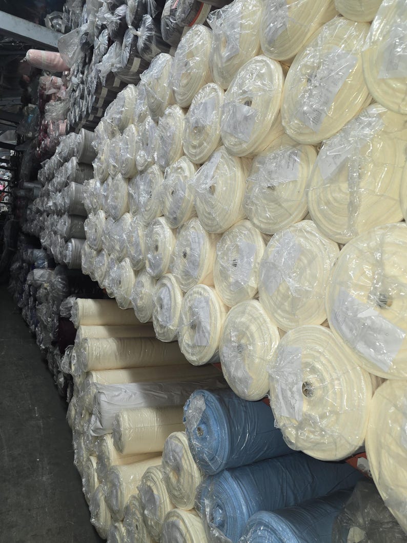 Puede incluir: Una gran pila de rollos de tela en varios colores, incluyendo blanco, beige y azul. Los rollos est&aacute;n envueltos en pl&aacute;stico transparente y apilados en un almac&eacute;n.