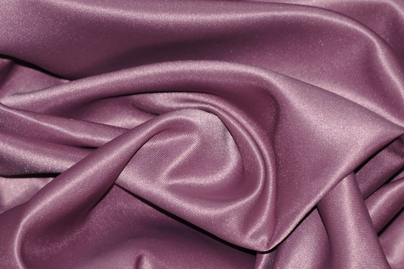 Puede incluir: Primer plano de una tela suave, lisa y malva con un brillo sutil. La tela est&aacute; drapeada y doblada, creando una textura lujosa y elegante.