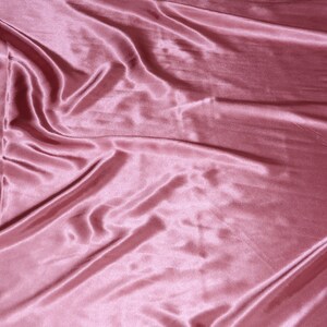 May include: A smooth, pink satin fabric with soft, flowing folds. The fabric has a subtle sheen and a luxurious feel.