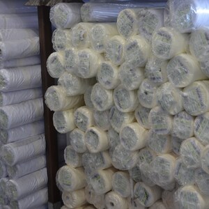 May include: Rolls of white fabric wrapped in clear plastic, stacked on shelves in a warehouse.