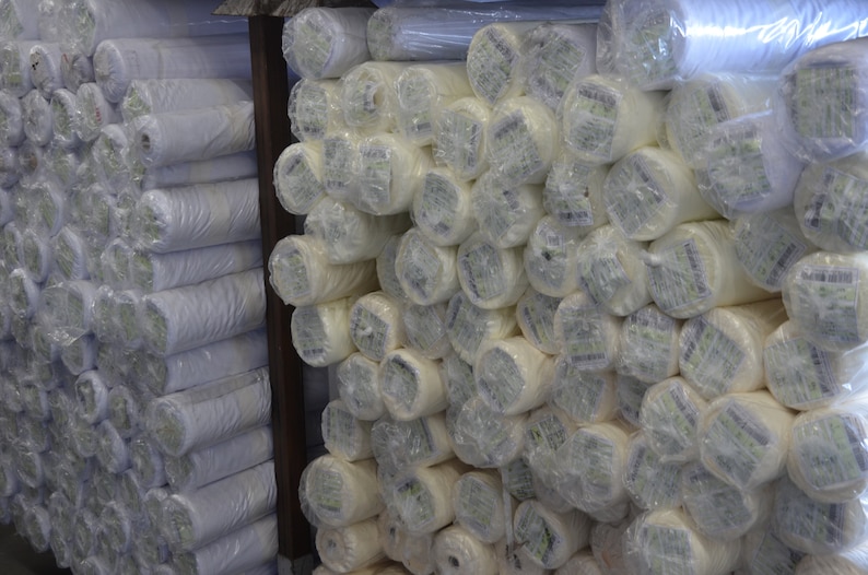 May include: Rolls of white fabric wrapped in clear plastic, stacked on shelves in a warehouse.