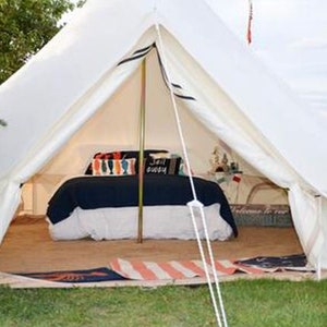 May include: A white canvas bell tent with a bed inside. The bed has a navy blue comforter and white pillows with colorful patterns. The tent has a wooden floor and a rug.