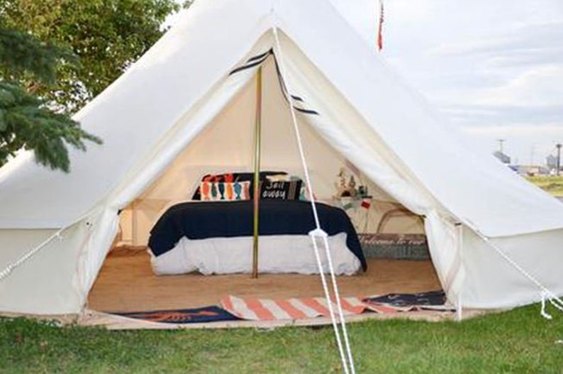 May include: A white canvas bell tent with a bed inside. The bed has a navy blue comforter and white pillows with colorful patterns. The tent has a wooden floor and a rug.