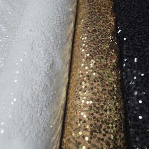 May include: Three rolls of fabric with sequins. The fabric on the left is white, the middle fabric is gold, and the fabric on the right is black.