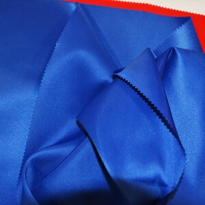 Puede incluir: Primer plano de una tela de sat&eacute;n azul doblada y drapeada sobre una superficie roja. La tela tiene una textura suave y sedosa.