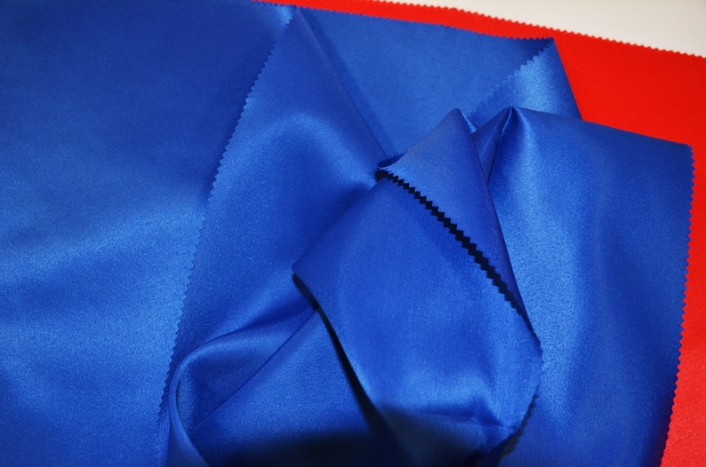 Puede incluir: Primer plano de una tela de sat&eacute;n azul doblada y drapeada sobre una superficie roja. La tela tiene una textura suave y sedosa.