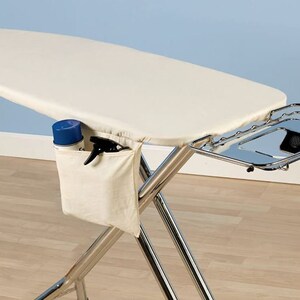 May include: A white ironing board with a chrome frame and a metal tray. The ironing board has a white fabric cover and a pocket on the side for storing ironing supplies. The tray is attached to the side of the ironing board and can be used to hold items like a spray bottle or iron.