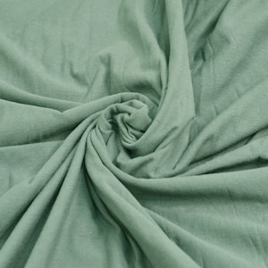 May include: A close-up of a soft, sage green fabric with a smooth texture. The fabric is folded and draped, creating a swirling pattern.