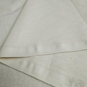 May include: Close-up of a plain, off-white fabric with a subtle texture. The fabric is folded in half, showing the edges of the material.