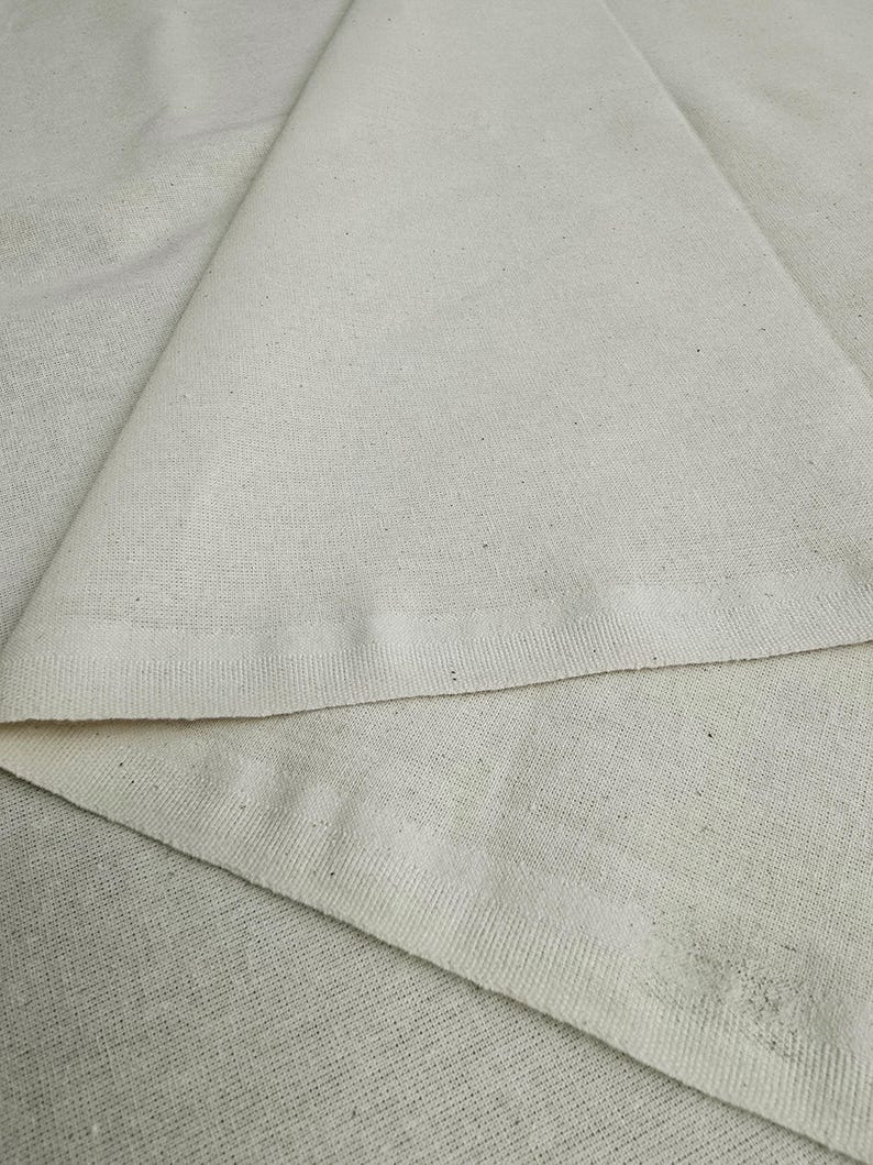 May include: Close-up of a plain, off-white fabric with a subtle texture. The fabric is folded in half, showing the edges of the material.