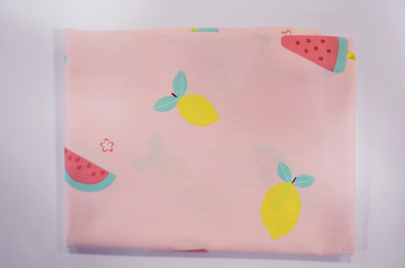 May include: Pink fabric with a repeating pattern of lemons and watermelon slices. The lemons are yellow with green leaves and the watermelon slices are red with black seeds and green rinds.