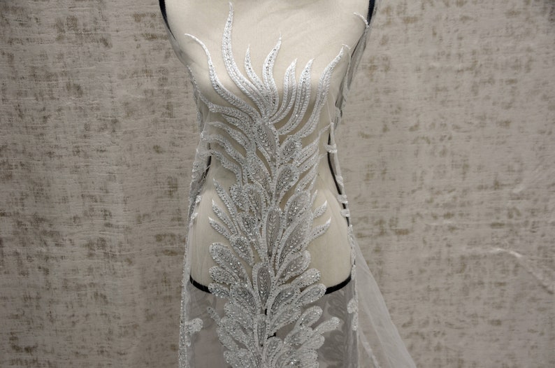 Puede incluir: Tela de encaje blanca con cuentas con un delicado patr&oacute;n floral. La tela es transparente y tiene una textura suave y fluida.