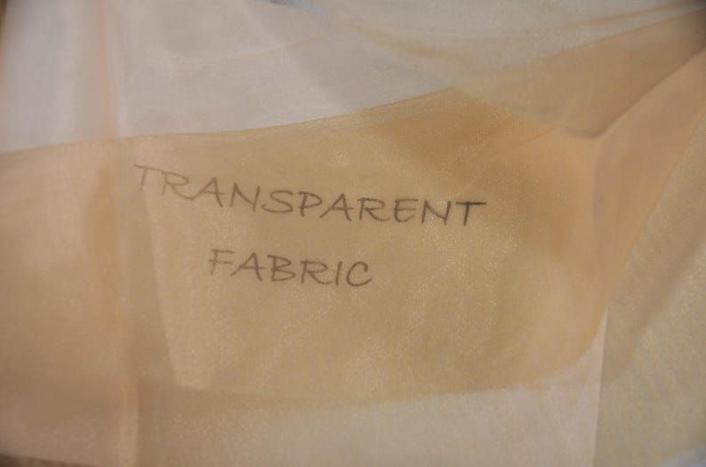 May include: A close-up of a piece of sheer, beige fabric with the words "TRANSPARENT FABRIC" written on it.