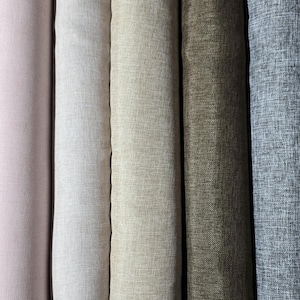 May include: A close-up of several rolls of fabric in various neutral colors. The colors range from a soft pink to a light gray-blue, with shades of white, beige, and olive green in between. The fabric has a textured appearance.