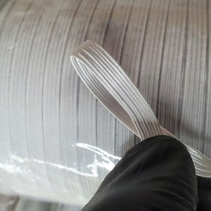 May include: A spool of white elastic cord, wound tightly and wrapped in clear plastic. The cord is about 1/4 inch wide and has a smooth, slightly textured surface.