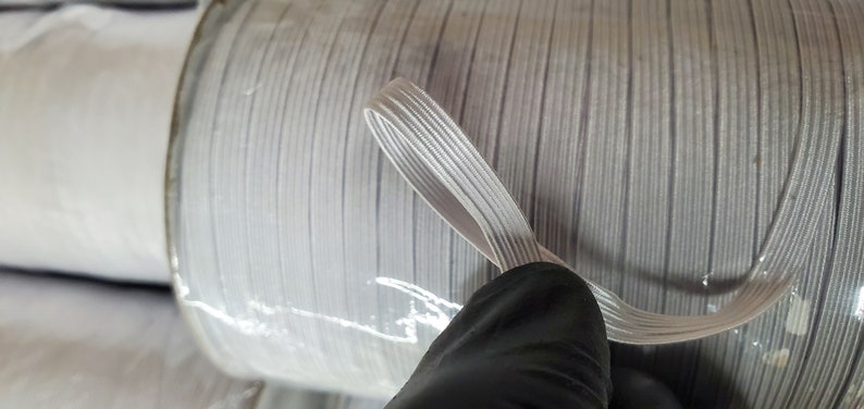 May include: A spool of white elastic cord, wound tightly and wrapped in clear plastic. The cord is about 1/4 inch wide and has a smooth, slightly textured surface.
