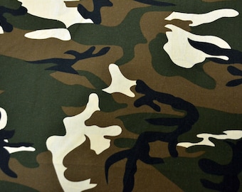 May include: Camouflage fabric with a green, brown, black, and white pattern. The fabric is shown next to a ruler.