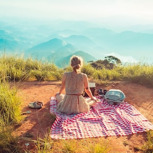 May include: A woman sits on a red and white checkered picnic blanket on a grassy hilltop overlooking a mountain range.