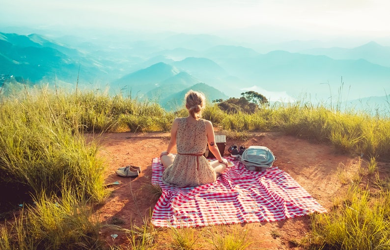 May include: A woman sits on a red and white checkered picnic blanket on a grassy hilltop overlooking a mountain range.