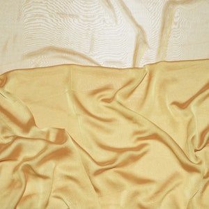 May include: A close-up of a gold fabric with a smooth, silky texture. The fabric is draped and folded, creating soft, flowing lines.