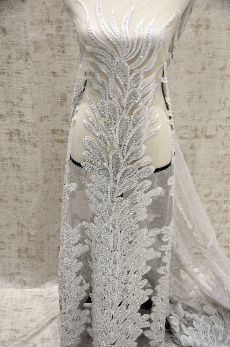 Puede incluir: Tela de encaje plateada con cuentas y un delicado patr&oacute;n floral. La tela es transparente y tiene una textura suave y fluida.