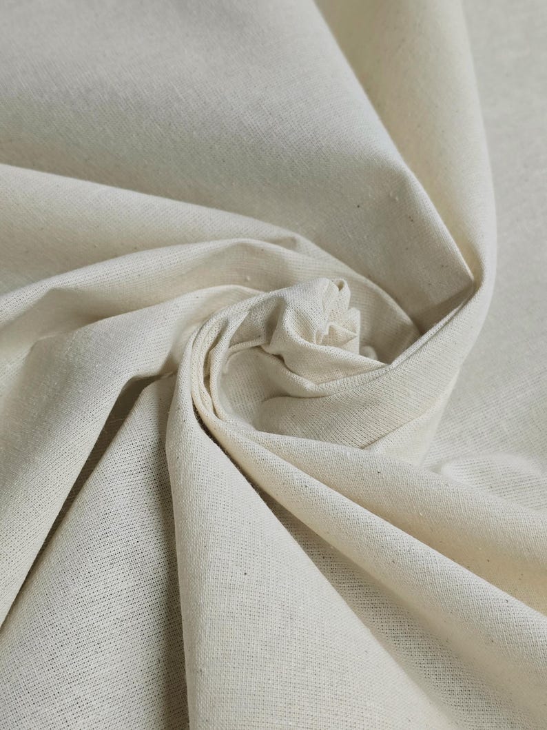 May include: Close-up of a natural, off-white fabric with a smooth, woven texture. The fabric is folded and draped, showcasing its soft and pliable nature.