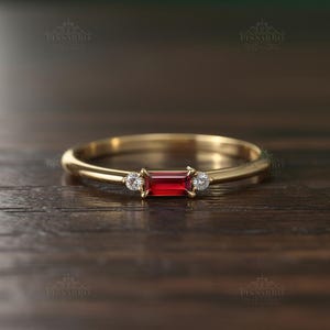 May include: A gold ring featuring a rectangular, vibrant red gemstone flanked by two smaller, round, clear gemstones. The ring has a thin band and is set against a dark wooden surface, highlighting the ring's design.