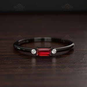 May include: A black ring with a rectangular, red gemstone at the center, flanked by two smaller, round, white gemstones. The ring has a sleek, minimalist design, perfect for everyday wear. The ring is set against a dark wooden surface.