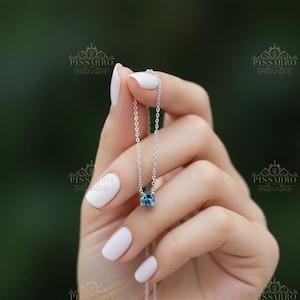 May include: A silver necklace with a round, blue gemstone pendant. The necklace is held by a hand with white nail polish against a blurred green background. The pendant is approximately 0.25 inches in diameter.
