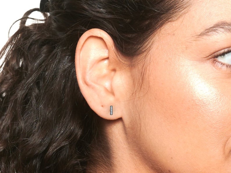 May include: A close-up of a person's ear with a small, silver bar earring with black diamonds. The earring is simple and elegant.