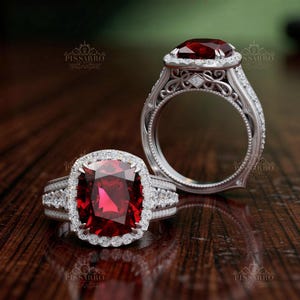 May include: Two ornate rings with large, square-cut, red gemstones. The rings are set in a silver metal with intricate detailing and surrounded by smaller, clear gemstones. The rings are displayed on a dark wooden surface.