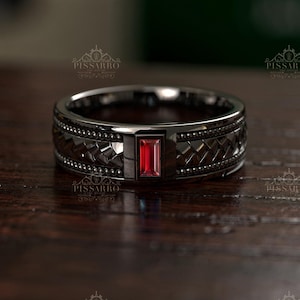 May include: A black ring with a rectangular red gemstone at the center. The ring has a textured band with a woven pattern and small black accents. The ring is displayed on a dark wooden surface.