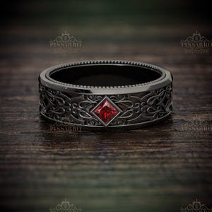 May include: A black ring with an ornate, swirling pattern and a square, red gemstone at the center. The ring has a textured band and a detailed design. The ring is displayed on a dark wooden surface.
