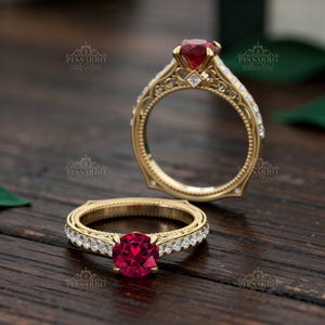 May include: Two gold rings with a ruby gemstone and small diamonds. The rings have an ornate, filigree design. The ruby is a deep red color. The rings are on a wooden surface.