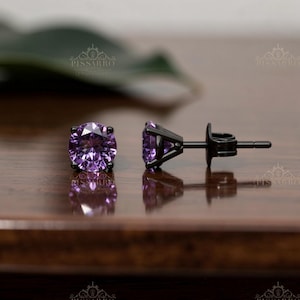May include: A pair of stud earrings featuring round, faceted purple gemstones set in black metal. The earrings are displayed on a wooden surface, with a blurred green leaf in the background. The earrings are a classic jewelry item.
