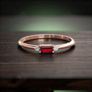 Peut inclure: Bague en or rose ornée d'une pierre rouge foncé rectangulaire, flanquée de deux petites pierres rondes et transparentes. La bague est posée sur une surface en bois sombre, mettant en valeur son design élégant et sa couleur vibrante.