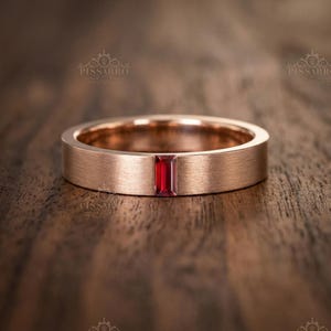 Peut inclure: Une bague en or rose avec une pierre précieuse rouge vif de forme rectangulaire au centre. La bande a une finition brossée, et la bague est présentée sur une surface en bois. La bague est un bijou.