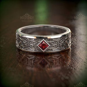 May include: A silver ring with an ornate, engraved design and a square, red gemstone at the center. The ring features a detailed pattern around the band and a textured edge. The ring is displayed on a dark wooden surface.