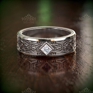 May include: A silver ring with an intricate floral design and a square-cut diamond at the center. The ring has a detailed pattern around the band and a textured edge. The ring is displayed on a dark wooden surface.