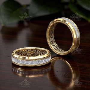 May include: Two gold wedding bands with intricate filigree designs. Each ring features a row of square-cut, clear gemstones set in a channel. The bands have a polished finish and are displayed on a dark wooden surface.