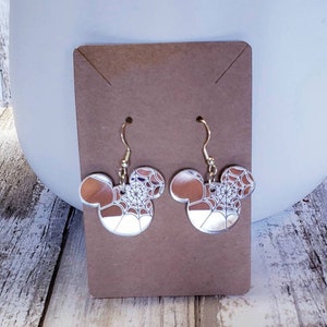 Op de afbeelding: Een paar zilveren oorhangers met een Mickey Mouse-silhouetontwerp. De oorbellen hebben een wit spinnenwebpatroon op een bruine achtergrond.