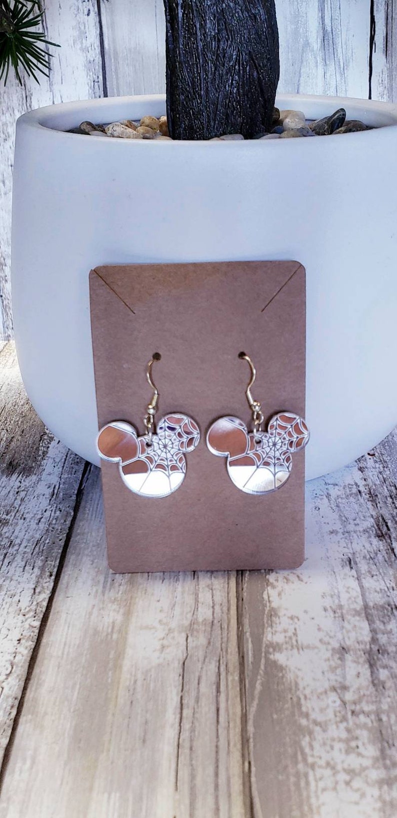 Op de afbeelding: Een paar zilveren oorhangers met een Mickey Mouse-silhouetontwerp. De oorbellen hebben een wit spinnenwebpatroon op een bruine achtergrond.