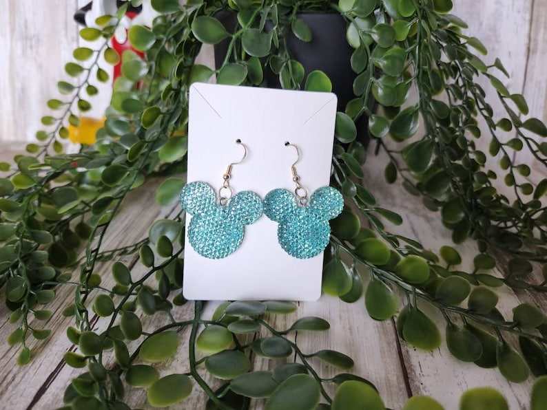May include: A pair of turquoise blue sparkly Mickey Mouse earrings. The earrings are on a white card.