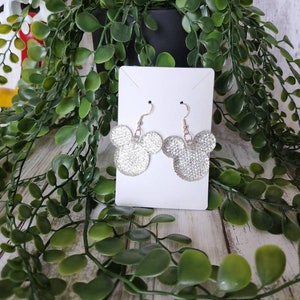 May include: Silver glitter Mickey Mouse earrings on a white card. The earrings are hanging from gold hooks.